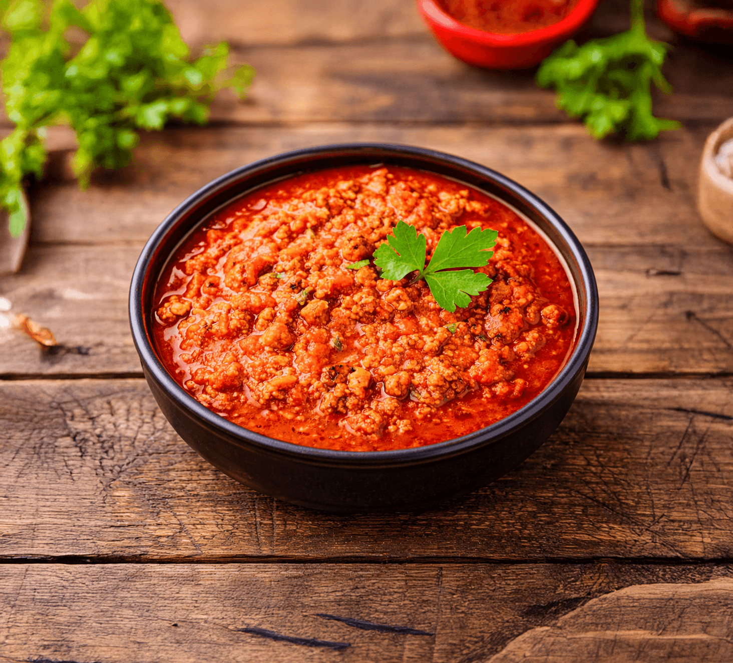 A bowl of tomato sauce on a wooden board with raw peppers, pasta, cheese, and herbs in a top-down rustic kitchen view.