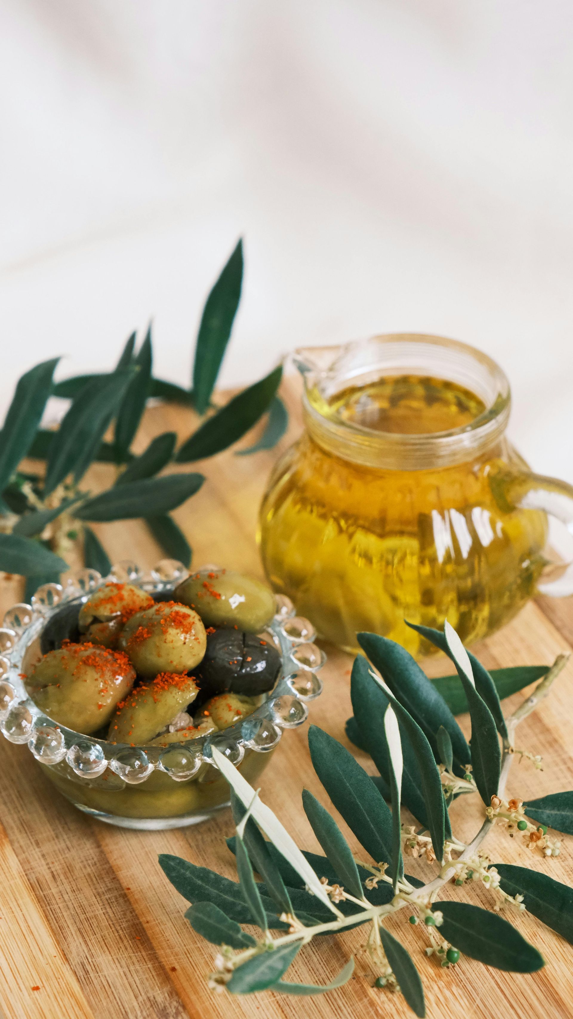 A glass bowl of seasoned olives beside a glass pitcher of olive oil and olive branches on a wooden board.