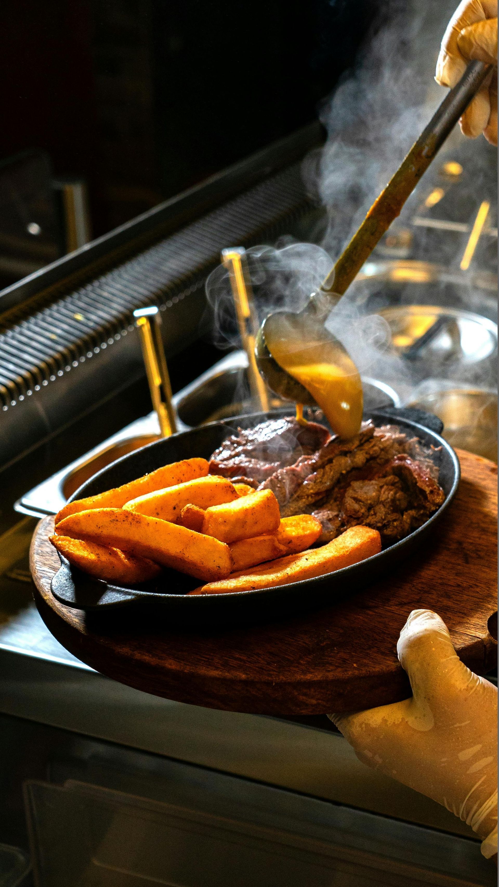 Sizzling skillet of steak and roasted vegetables on a table, with steam rising and a hand holding tongs