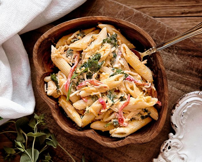 A bowl of creamy penne pasta mixed with sautéed greens and red bell pepper strips, set on a rustic wooden table.