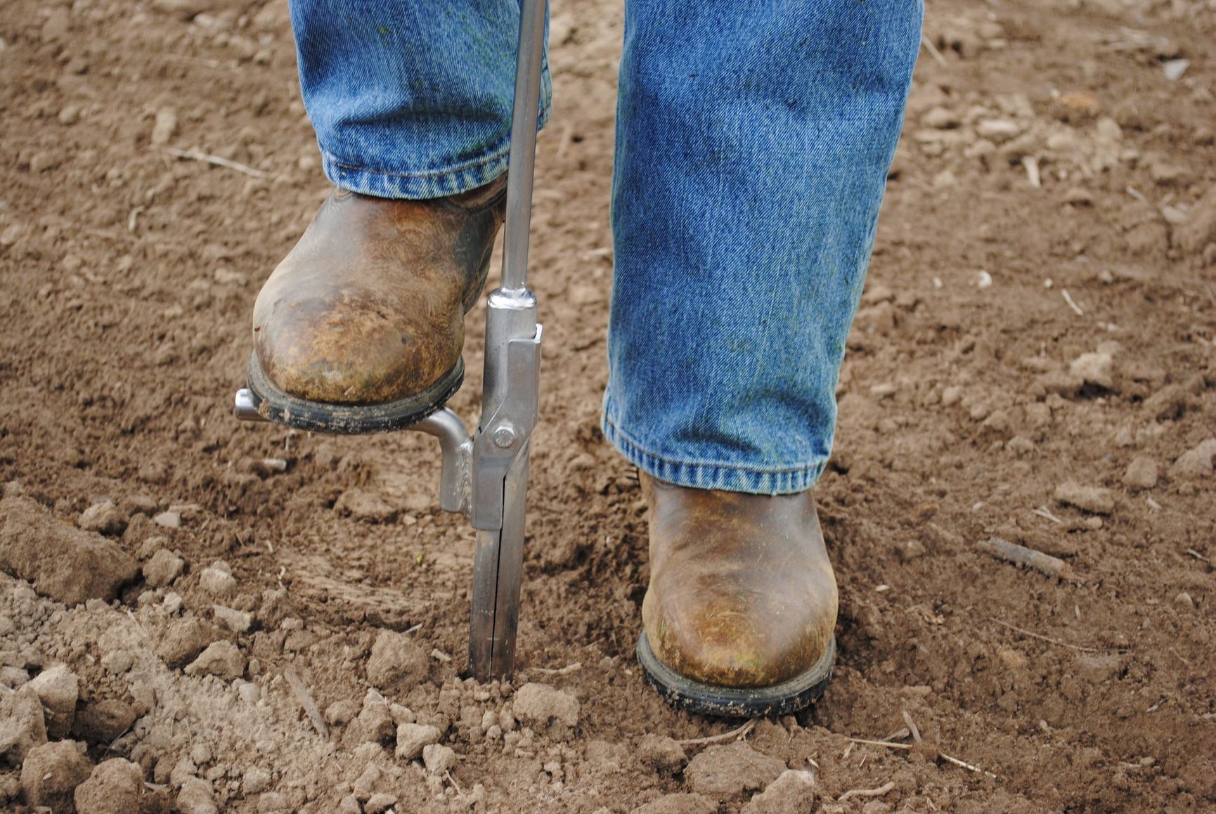 Booted feet standing on a post-hole digger in dirt soil