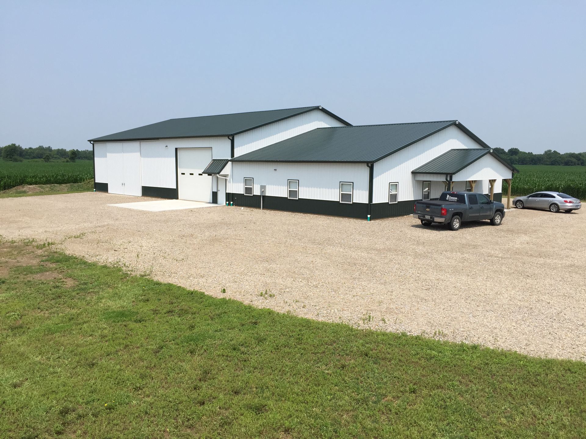 White metal building with green roof on gravel lot, with a truck and car parked outside.