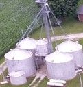 Aerial view of four white grain silos in a grassy field, linked by a central metal structure.