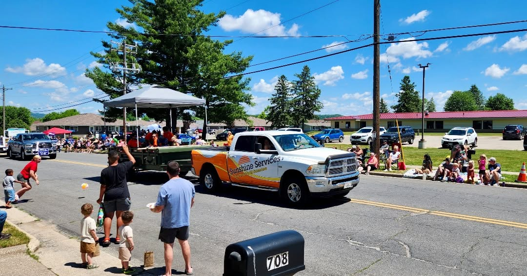 Parade truck decorated in orange and white drives past spectators on a sunny street with a gazebo and flags.