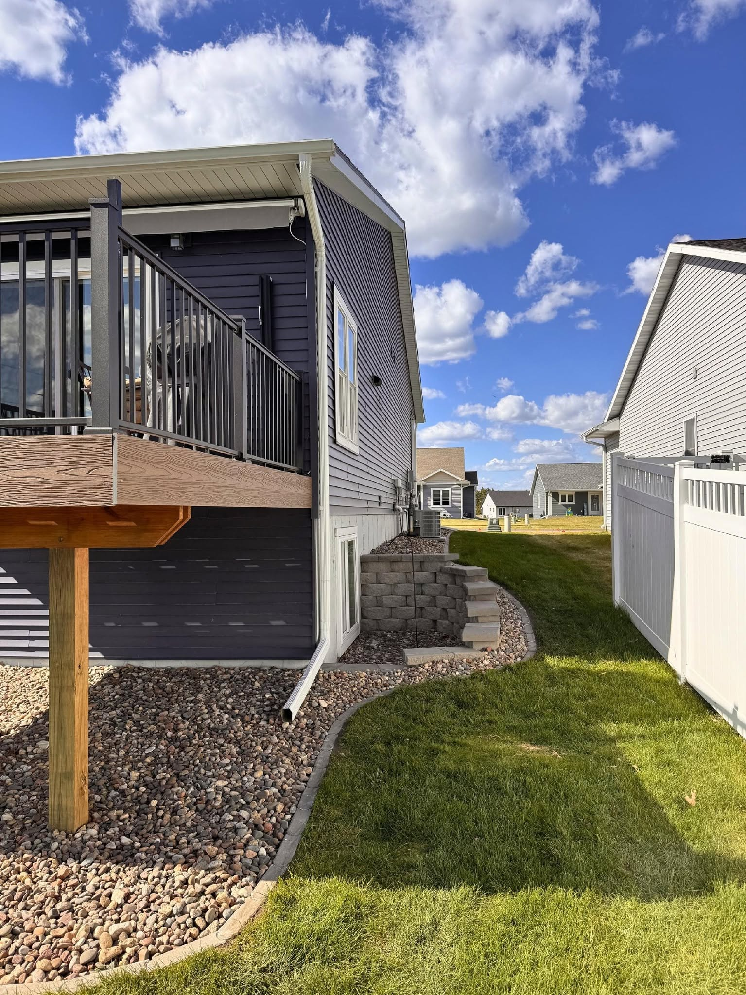 Backyard patio beside a white house with deck, stone edging, and green grass under a blue sky with clouds