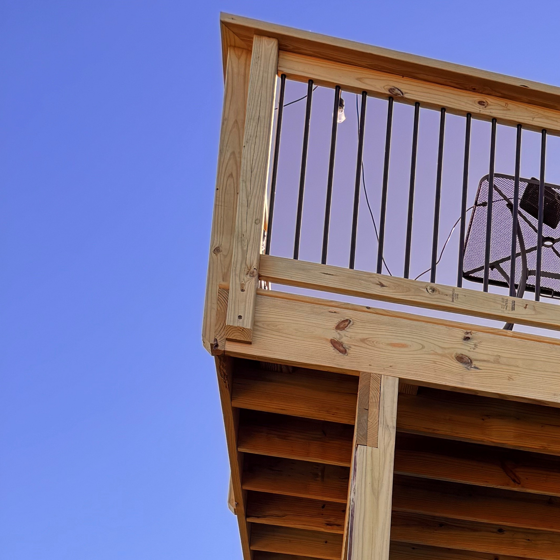 Wooden deck with black metal railing against a blue sky, an outdoor chair visible.