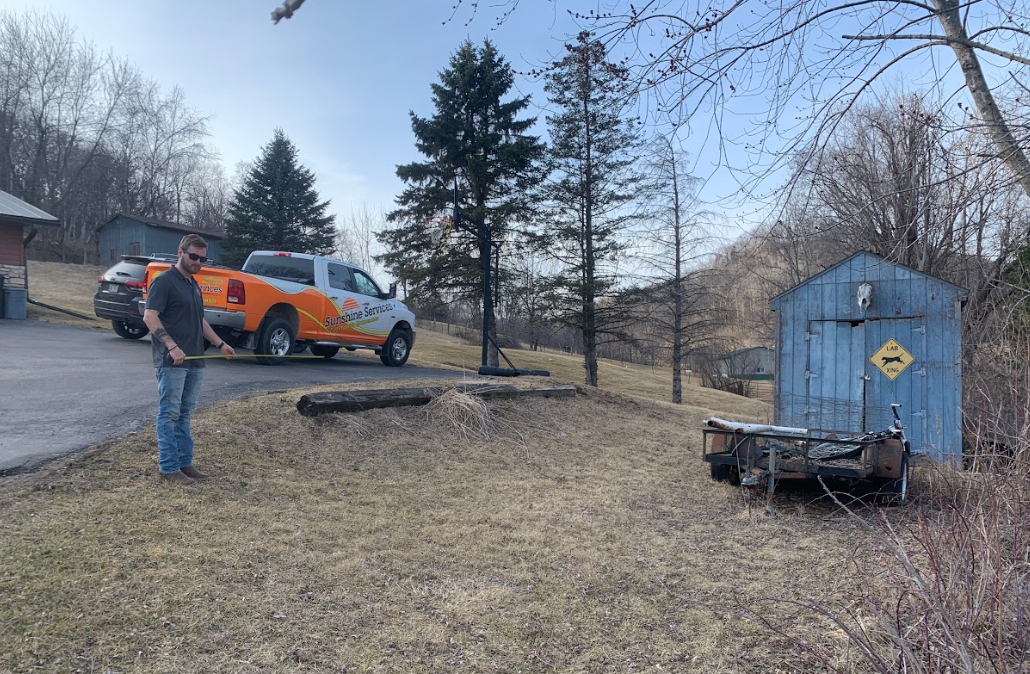 Man standing on grass, holding a tool, near an orange work truck and a blue shed in a yard.