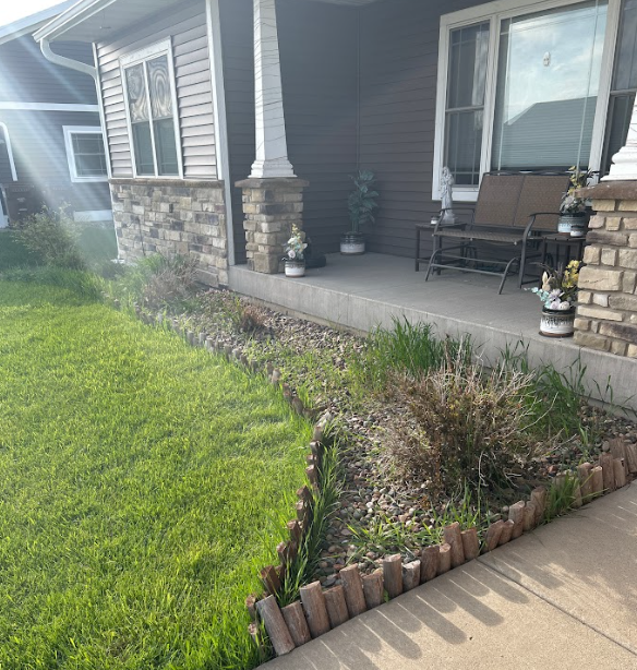 Front yard with a brown-sided house and porch. A flower bed with dead plants borders the porch, next to green grass.
