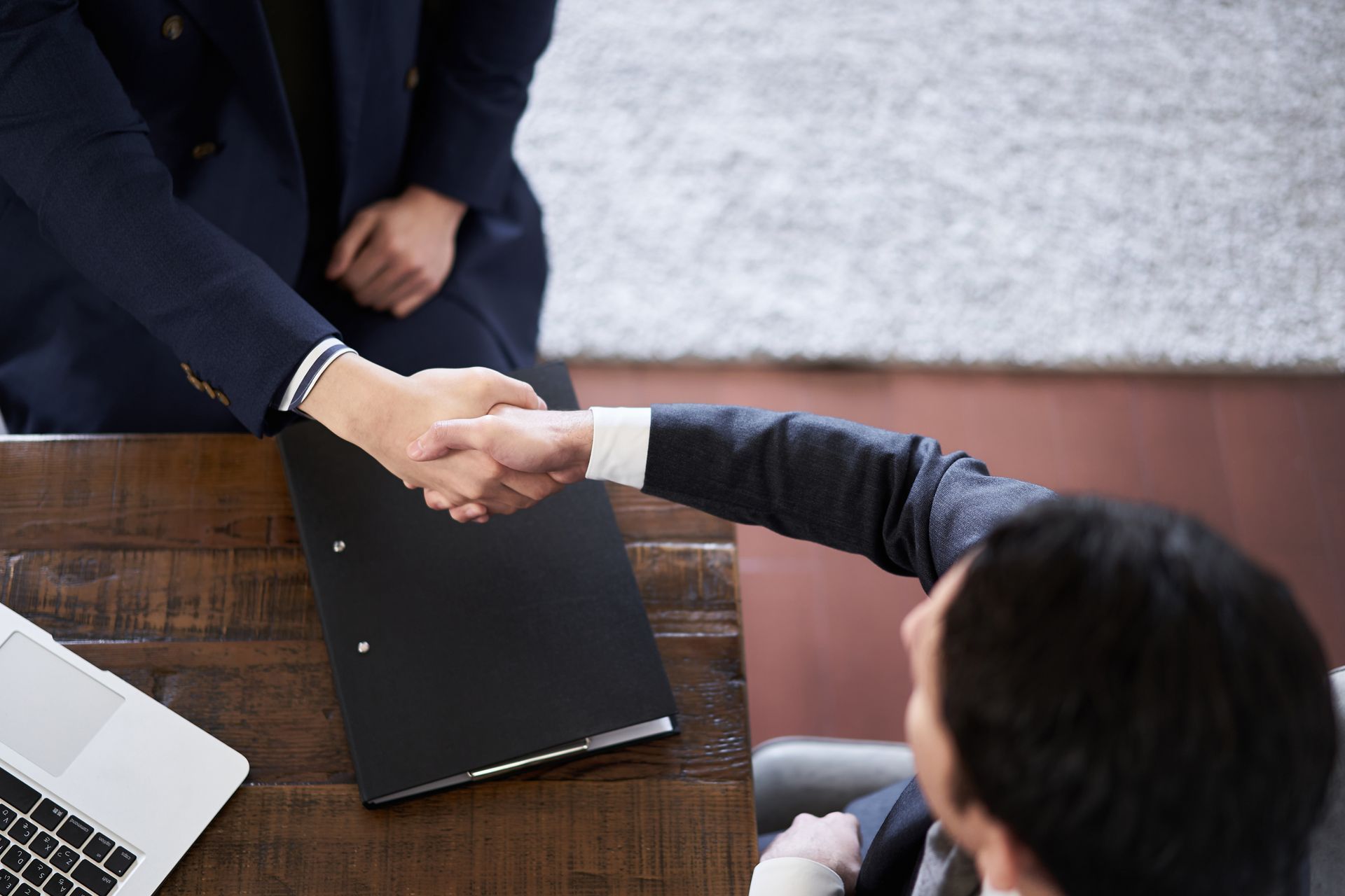 Two people in suits shaking hands over a wooden desk with a laptop and a black folder.