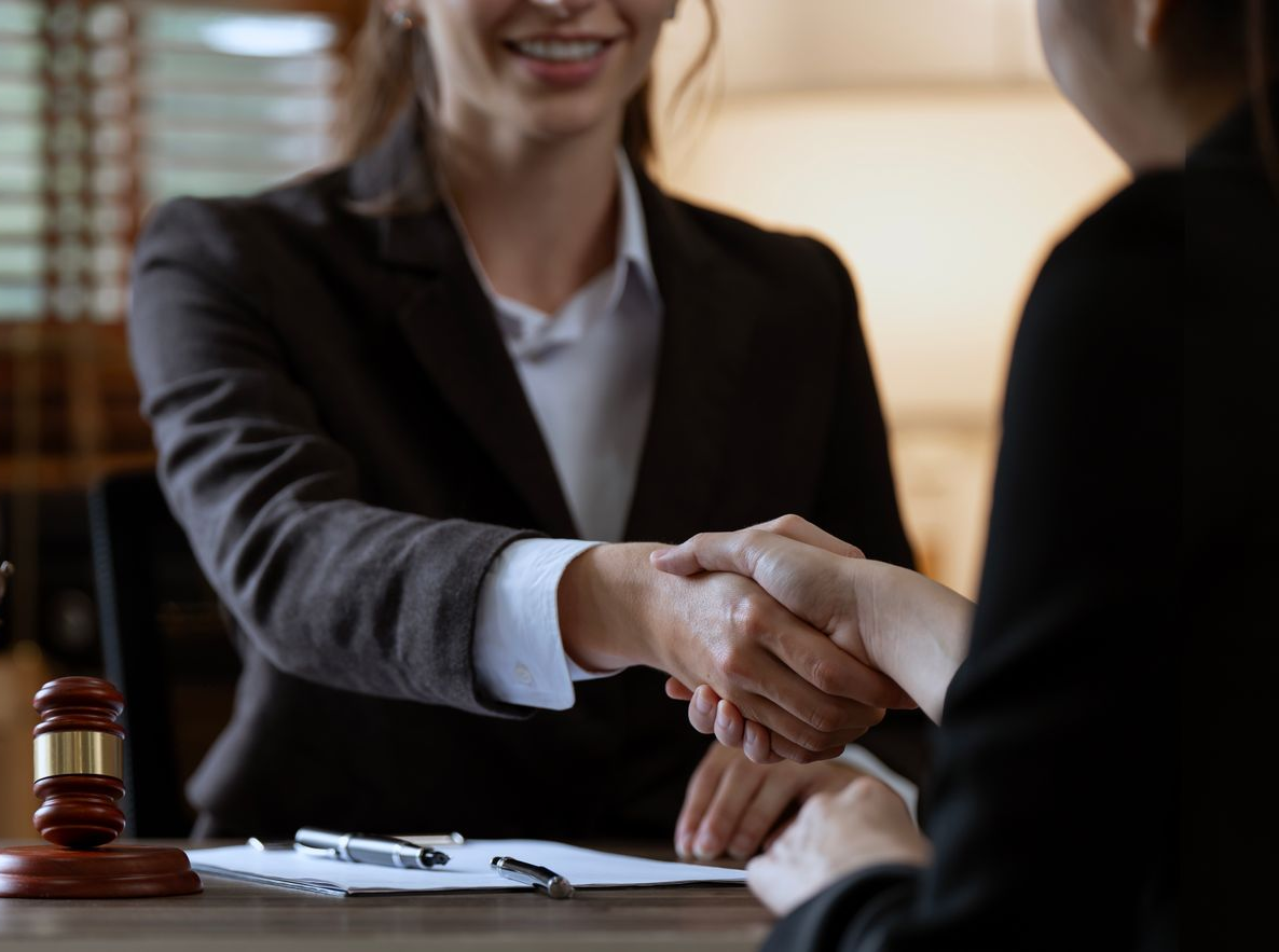 Two people in suits shaking hands in an office, gavel in foreground, smiling.