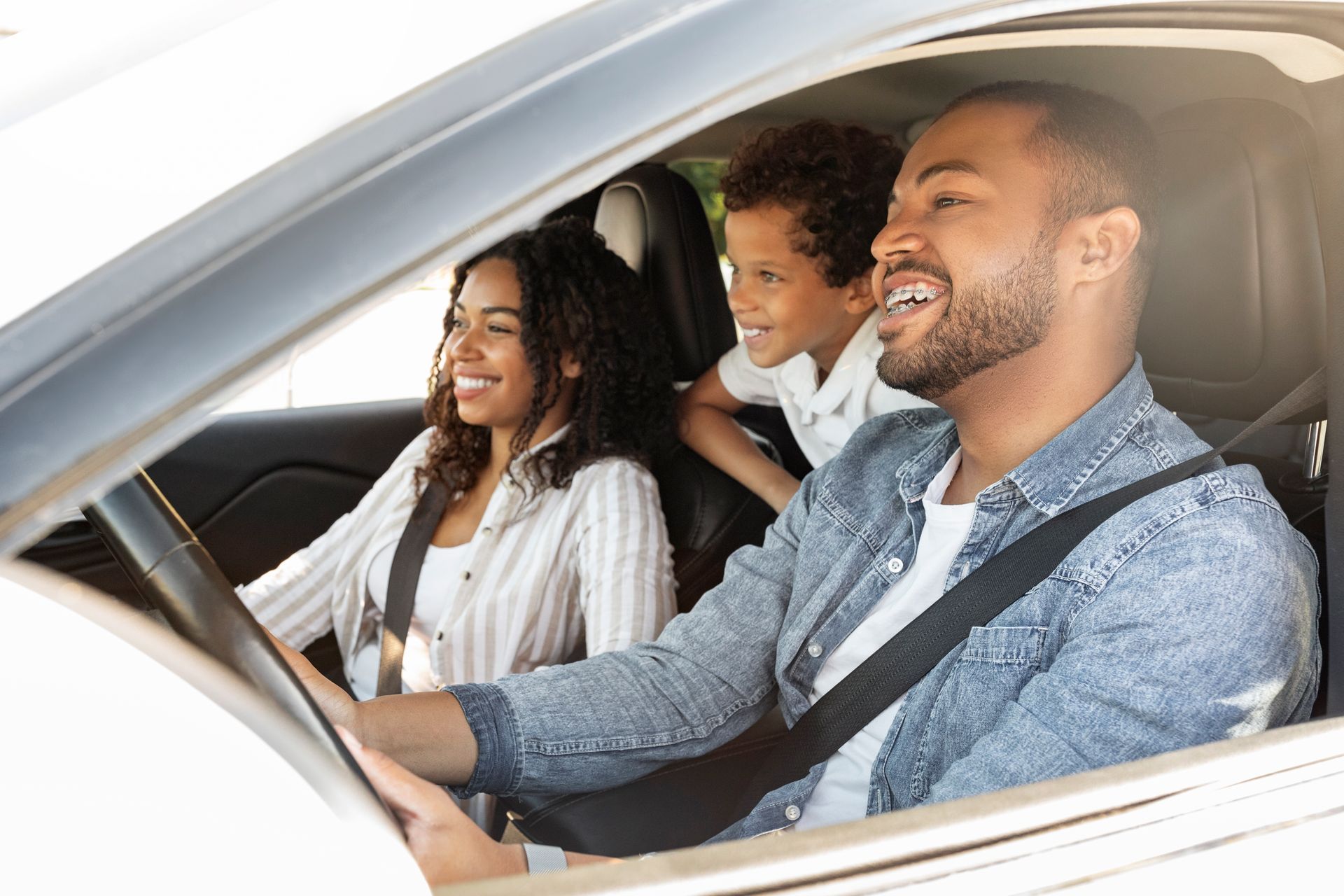 A family of three sitting inside a car, smiling and looking out the window while wearing seatbelts.