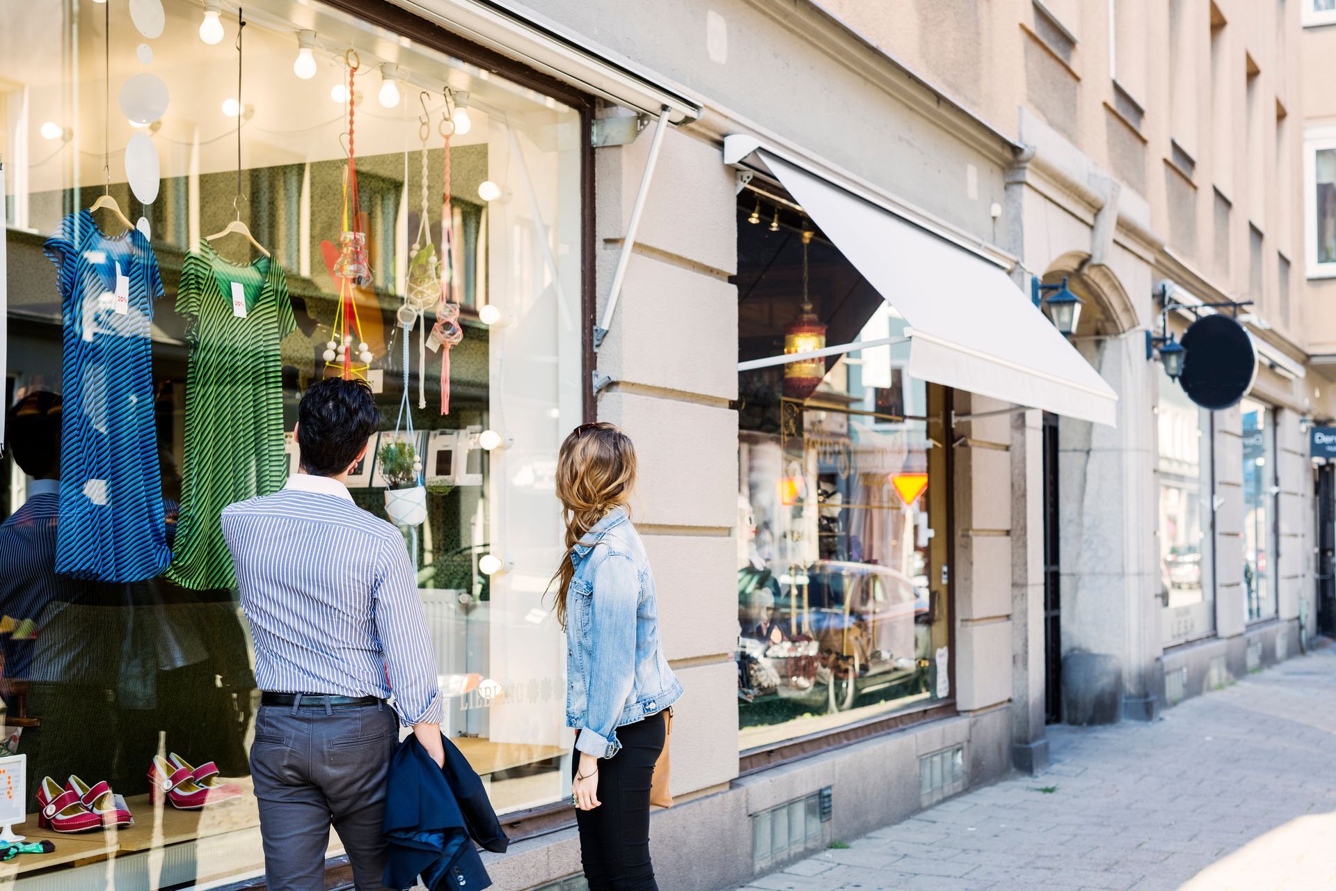 Two people stand on a sidewalk looking into a brightly lit retail store display window featuring colorful dresses.