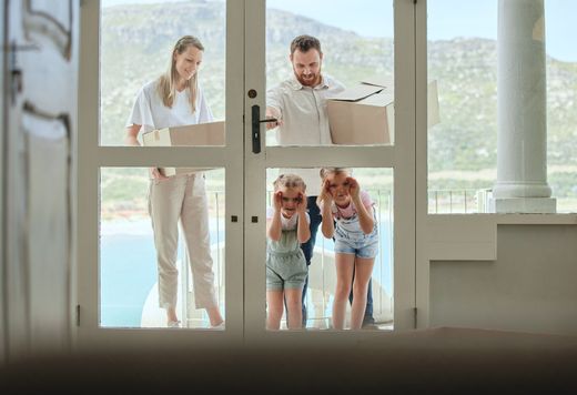 A family of four stands outside a glass door holding cardboard boxes, while the two children look inside through the glass.