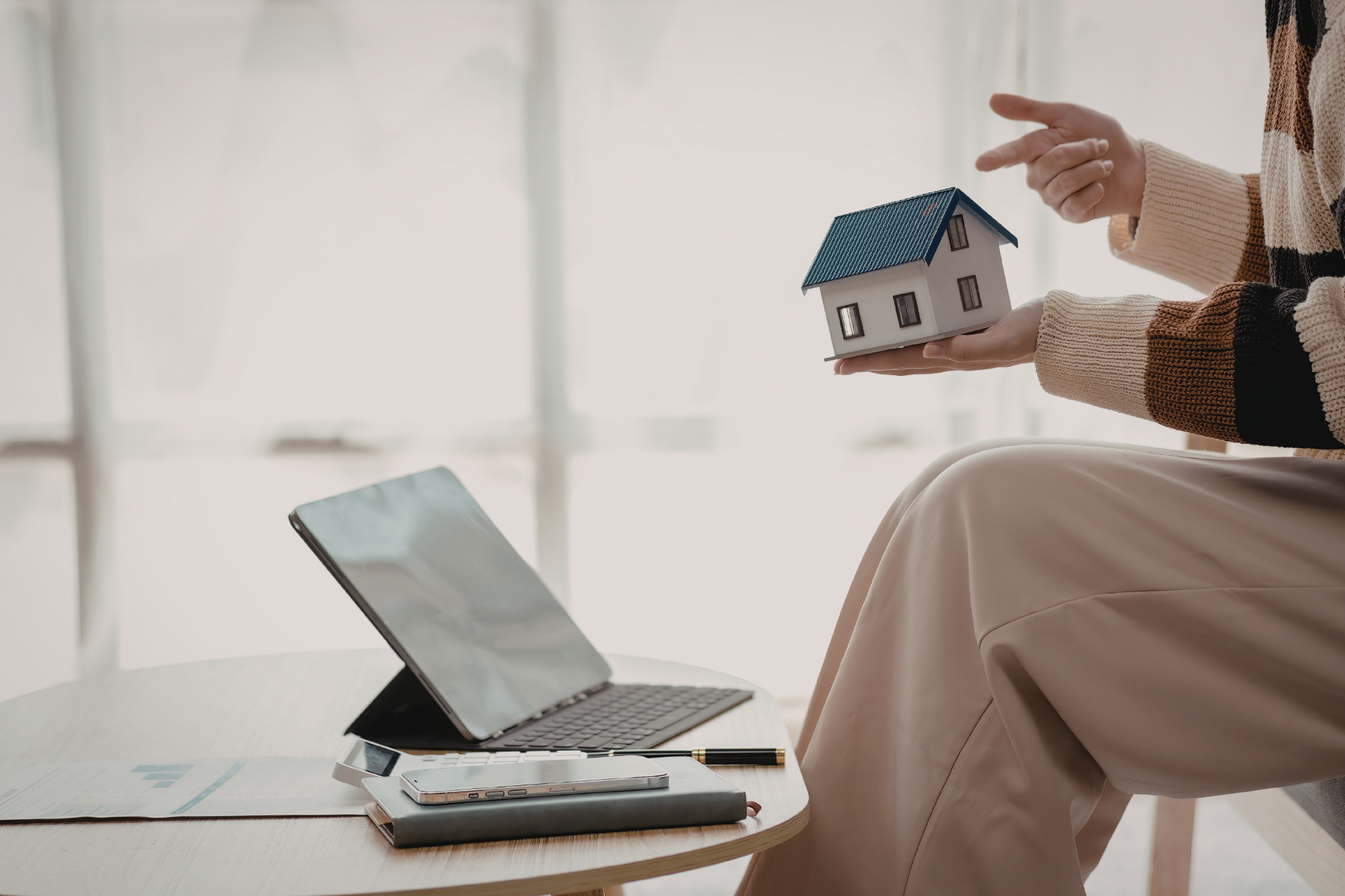 A person holds a small house model in their hand next to a laptop on a table, symbolizing real estate planning.