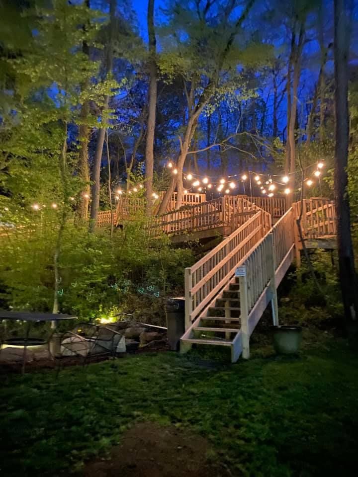 Wooden staircase with string lights leading up to a platform in a wooded area at dusk.