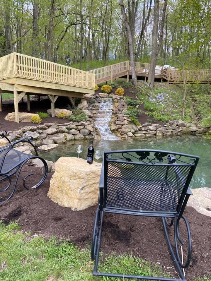 Black rocking chair by a pond with a small waterfall. Wooden bridge in background, trees surrounding.