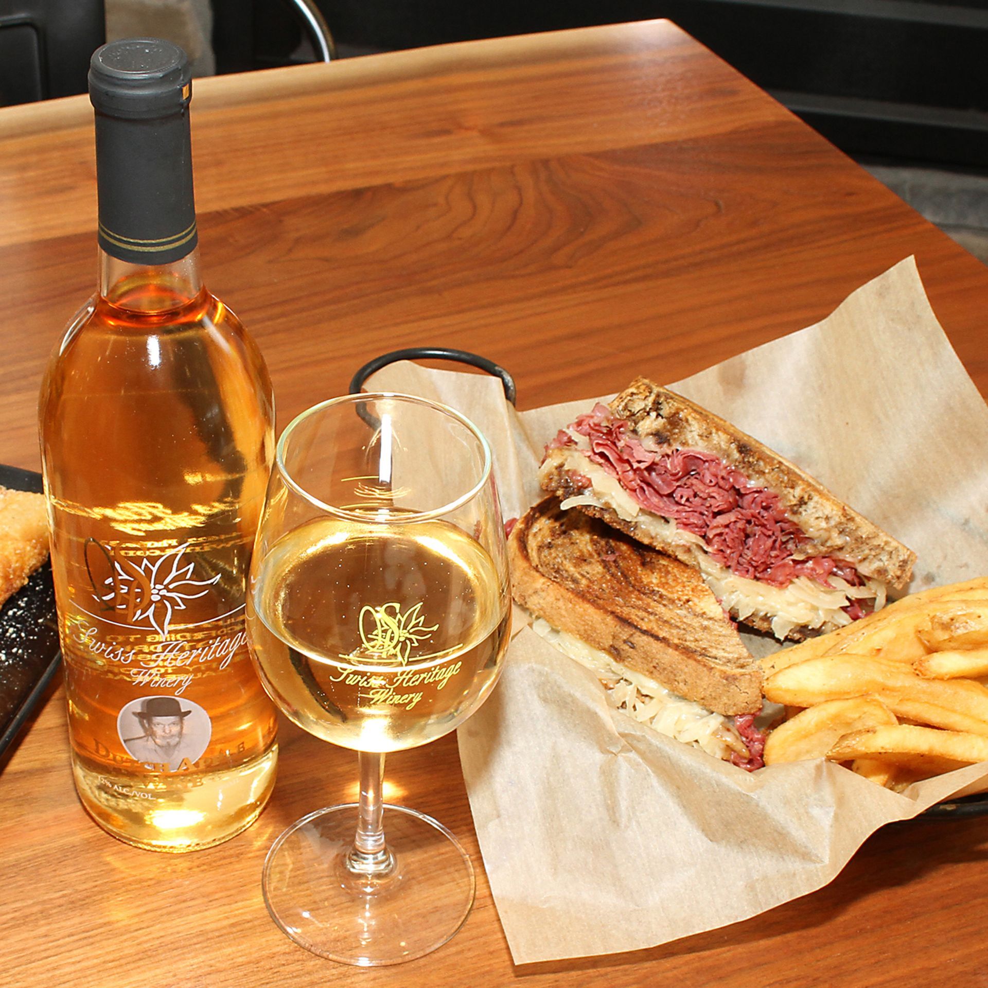 Bottle of wine, glass, and a Reuben sandwich with fries on a wooden table.