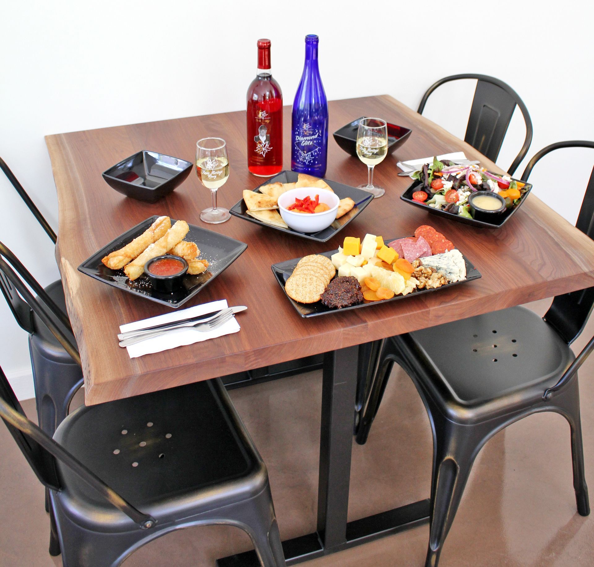 Wooden table with food, drinks, and black metal chairs. Includes wine bottles, charcuterie, and salads.