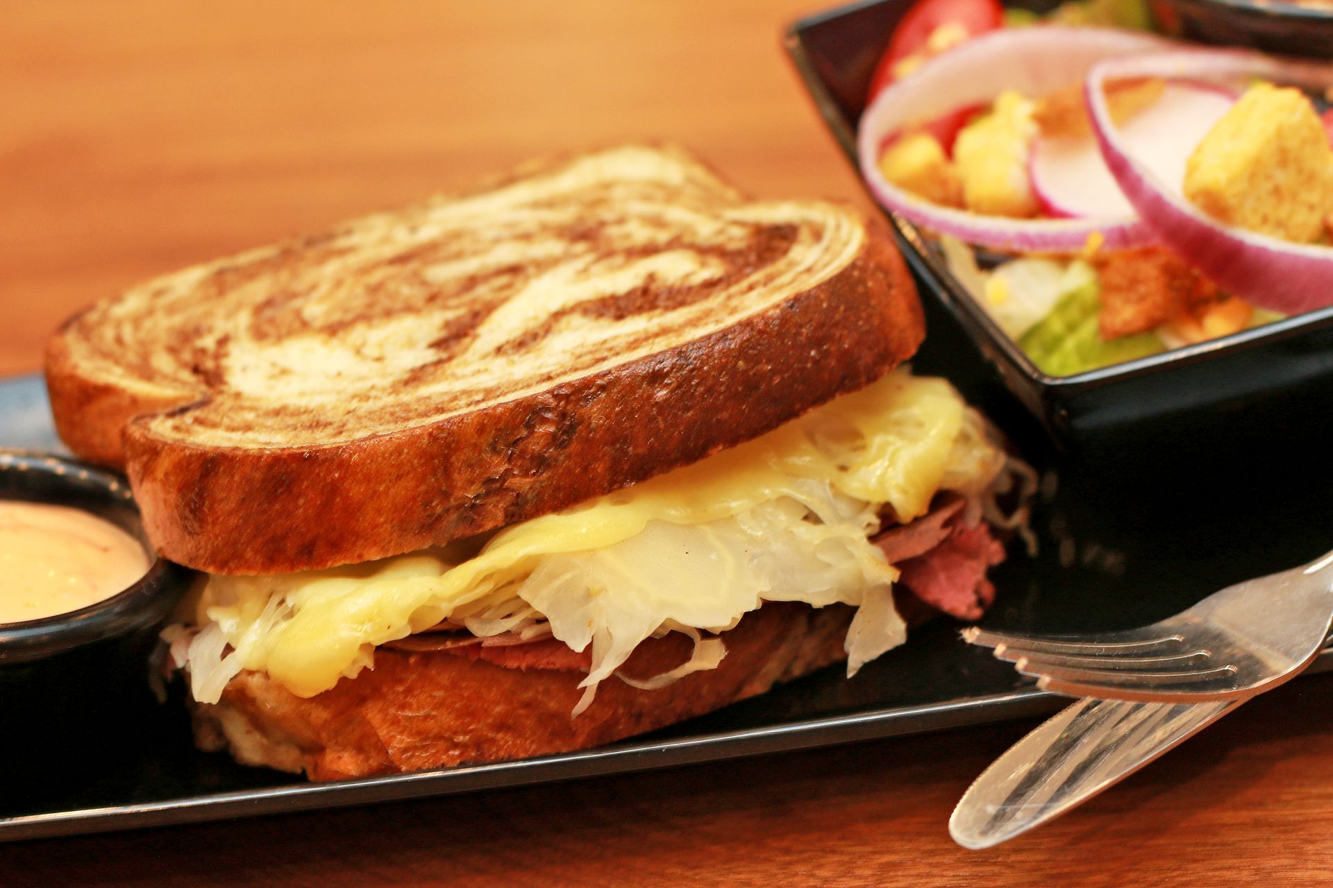 Reuben sandwich with melted cheese, sauerkraut, and a side salad on a black plate.