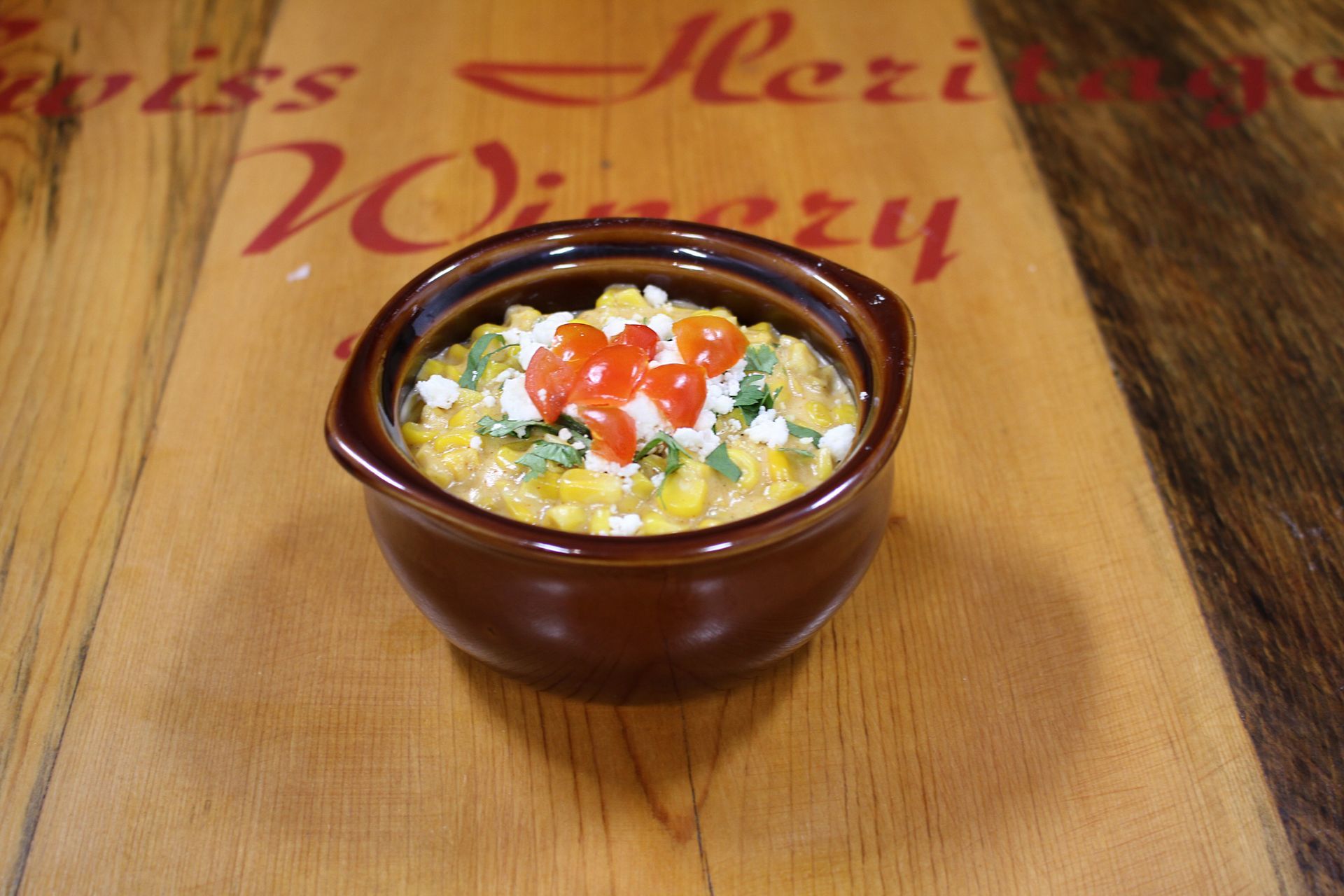 Corn dish in a brown bowl topped with tomatoes and herbs on a wooden board.