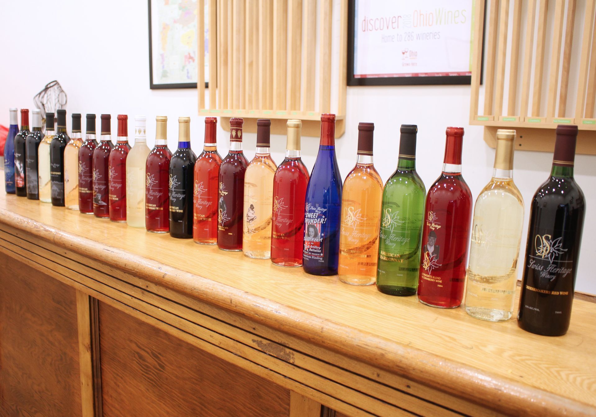 Bottles of various colored wines lined up on a wooden counter inside a room.