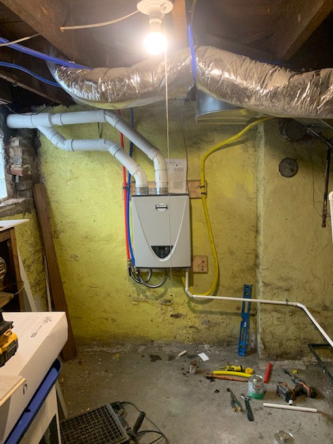 A tankless water heater mounted on a yellow basement wall, connected to white PVC vent pipes and a yellow gas line.