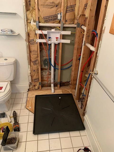 In-wall laundry plumbing hookups, white drainage pipe, and a black drip pan installed in a bathroom wall under renovation.