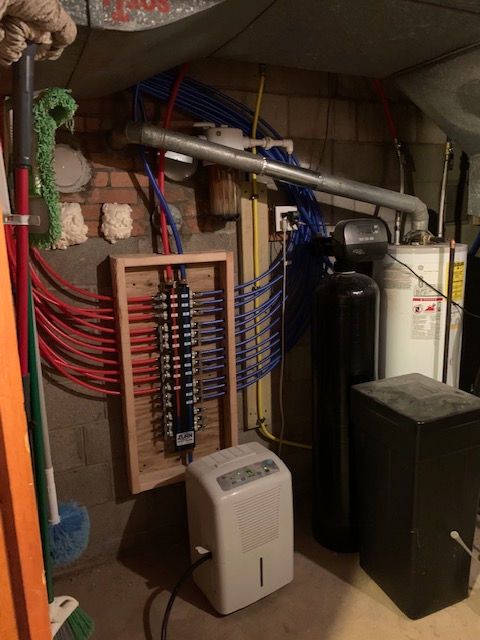 Basement utility area showing a red and blue PEX plumbing manifold mounted on a wooden frame with a water softener nearby.