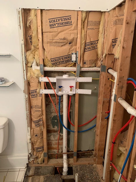 Wall cavity with exposed wooden studs, fiberglass insulation, and a new white washer outlet box with blue and red valves.