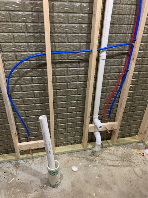 Unfinished basement wall with exposed wood studs, a vertical white PVC pipe, and blue and red PEX plumbing lines.