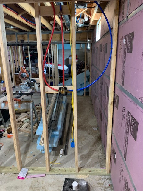 An unfinished basement wall frame with red and blue PEX plumbing pipes and electrical wiring installed.