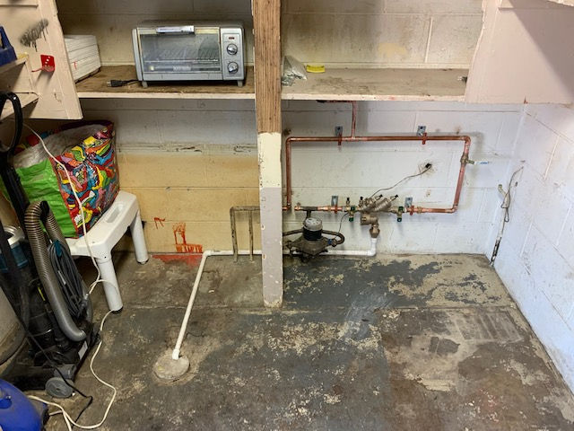Utility room corner with copper plumbing, a water meter on the floor, and a shelf holding a toaster oven above.