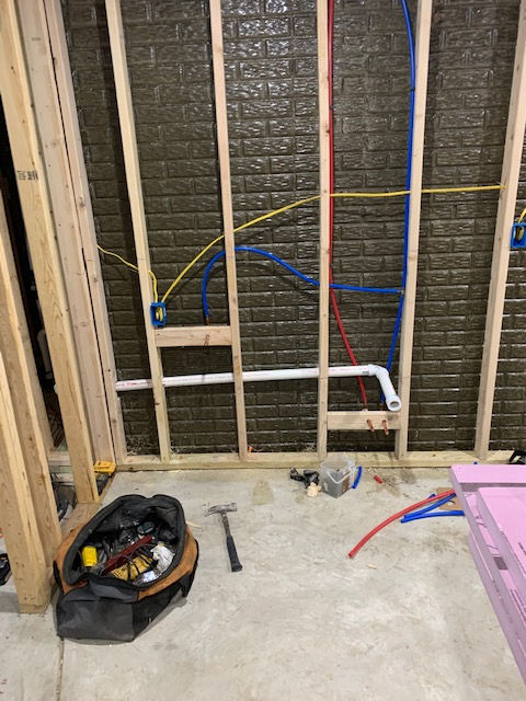 A partially framed basement wall with exposed white PVC plumbing pipes, blue and red PEX water lines, and yellow wiring.