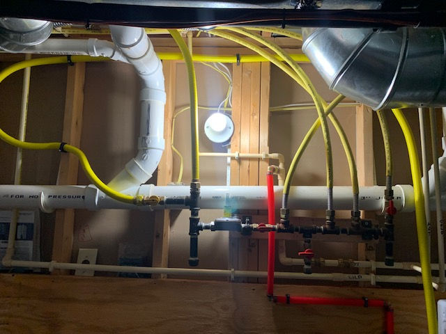 Exposed ceiling with yellow flexible gas pipes, white PVC plumbing, and red PEX water lines connected to a manifold.