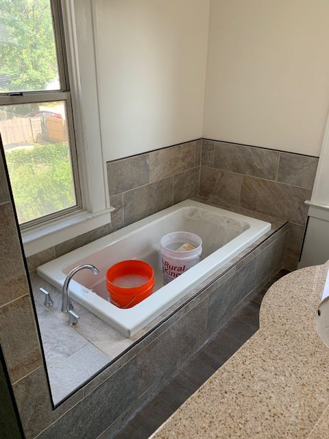 A white bathtub with an orange and a white bucket inside, set against tile walls in a sunlit bathroom.
