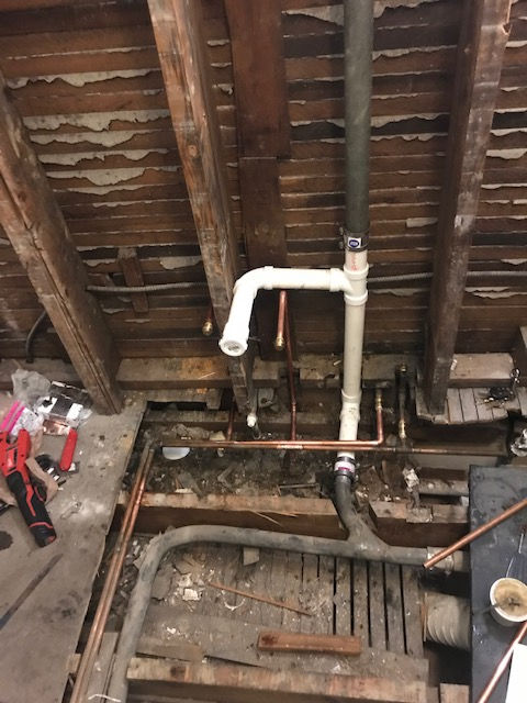 Construction scene showing new white PVC plumbing pipes connected to older metal pipes between exposed wooden joists.