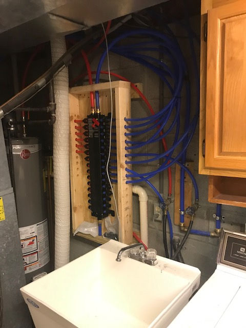 A plumbing manifold with blue and red PEX pipes mounted on a wooden frame above a utility sink in a basement.