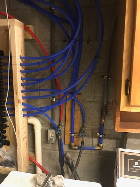 Blue PEX plumbing lines run across a cinderblock wall near a wooden mounting board and electrical outlet in a utility room.