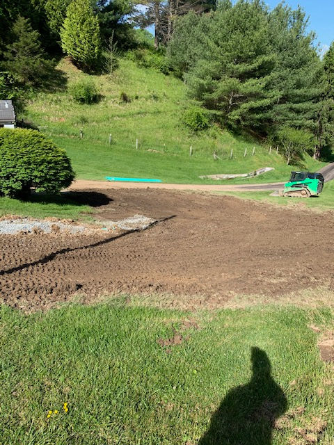 A dirt-covered area being prepared for landscaping, next to a green lawn, trees, and a small green construction machine.