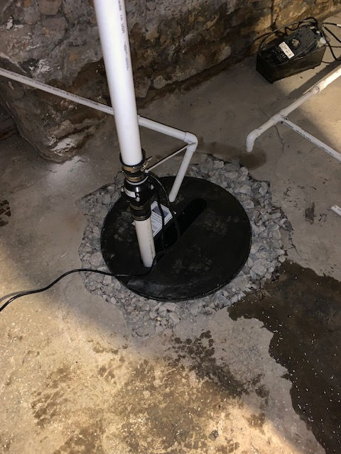 A sump pump installation in a basement floor with a vertical white PVC discharge pipe and a black circular basin cover.