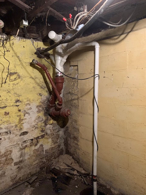 Plumbing pipes of PVC and red metal connected to a vertical drain pipe against a yellow block wall in a basement.