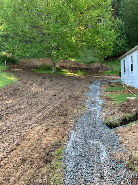 A gravel drainage trench runs along the side of a building next to a sloped, freshly graded dirt yard with a large tree.