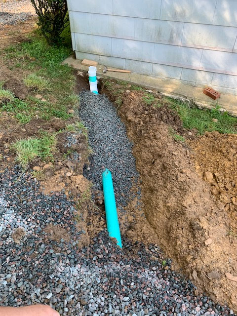 A trench with a blue pipe and gravel bedding running alongside a house foundation.