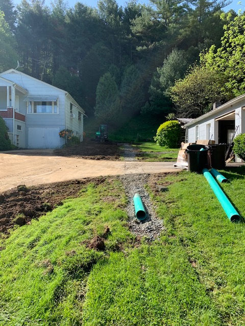 A green drainage pipe lies in a gravel trench on a grassy lawn between a house and a garage, with a dirt area nearby.