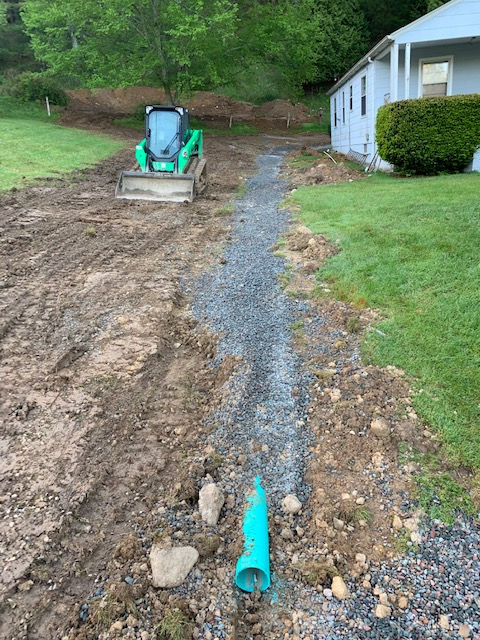 A green mini excavator sits in a muddy yard next to a gravel-filled trench with a protruding green drainage pipe.