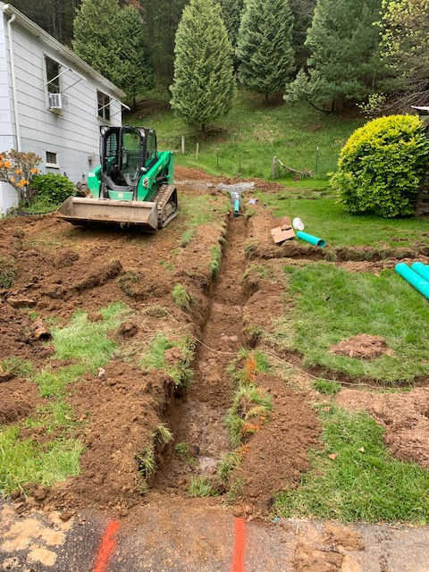 A green skid-steer loader sits beside a long, open trench in a grassy backyard near a white house.