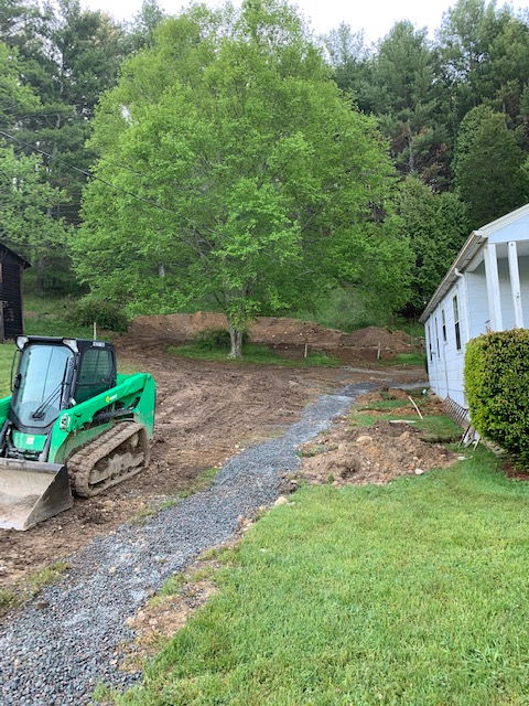 A green skid steer parked on a dirt lot next to a white house with a newly laid gravel path.