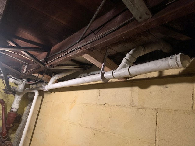 White PVC plumbing pipes run horizontally along a beige cinder block wall, suspended by metal hangers from floor joists.