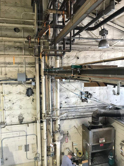 A high-angle view of an industrial space with overhead pipes, a metal railing, and a person working near machinery.