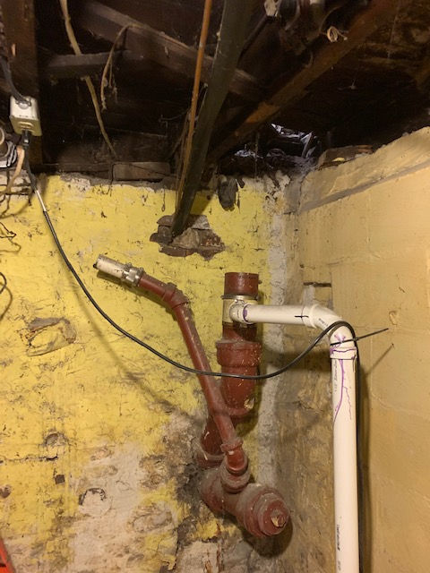 An interior basement corner showing cast iron and white PVC pipes attached to a yellow masonry wall with exposed wiring.